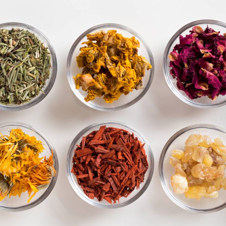 Six glass bowls filled with various dried herbs and flowers on a white background